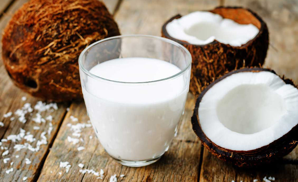 Glass of coconut milk on a wooden table, with actual coconuts around it 