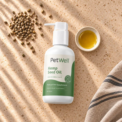 A bottle of PetWell Hemp Seed Oil pet supplement is displayed on a textured beige surface with dappled sunlight, alongside a small dish of golden oil, scattered hemp seeds, and a striped cloth.