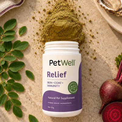 A white jar of PetWell Relief natural pet supplement with a purple label, styled on a beige stone surface with green leaves, a pile of green powder, and red beets.